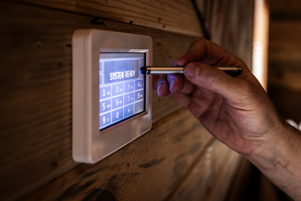Hand using a stylus to input a numeric code on a touch-screen commercial security panel mounted on a rustic wood plank wall, LCD reading SYSTEM READY
