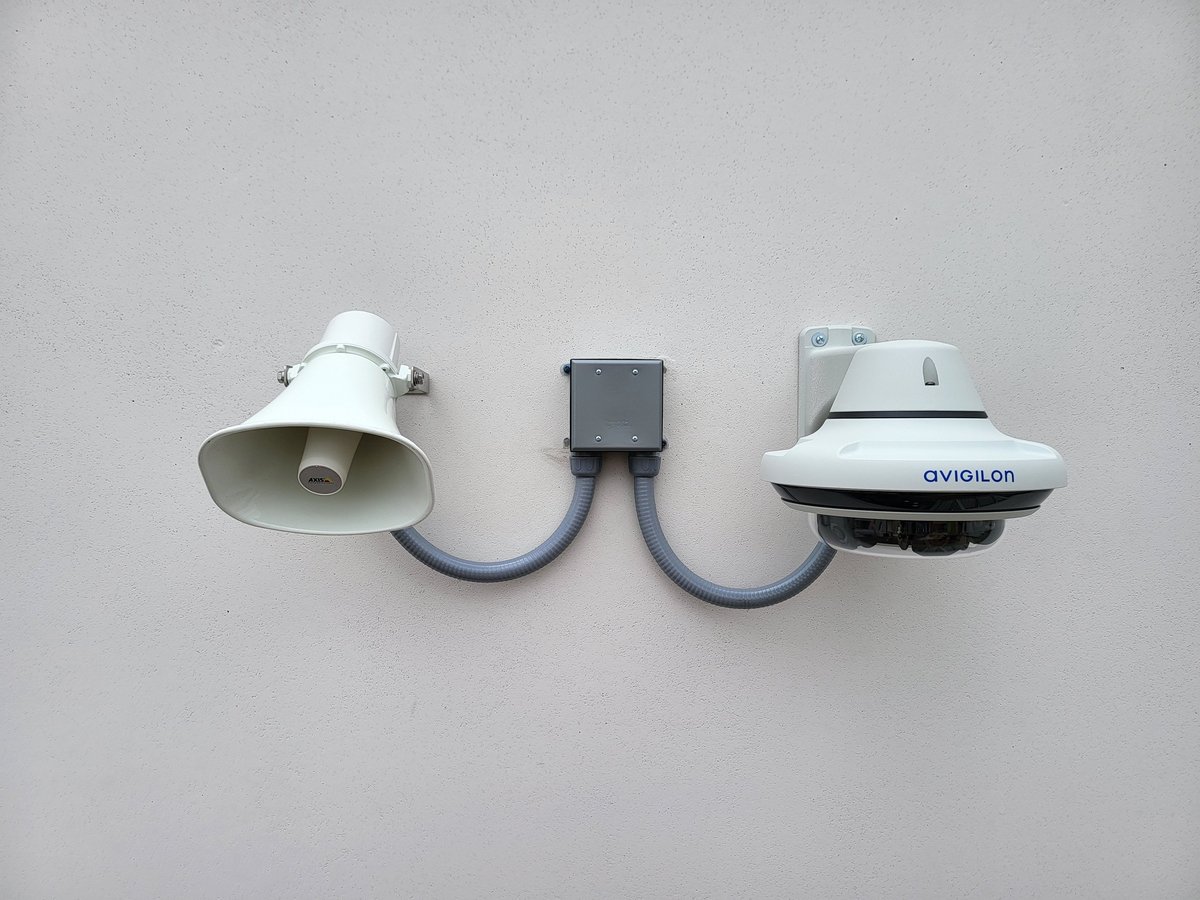 Two white security cameras connected by blue cables through a central gray junction box, mounted on a light gray wall