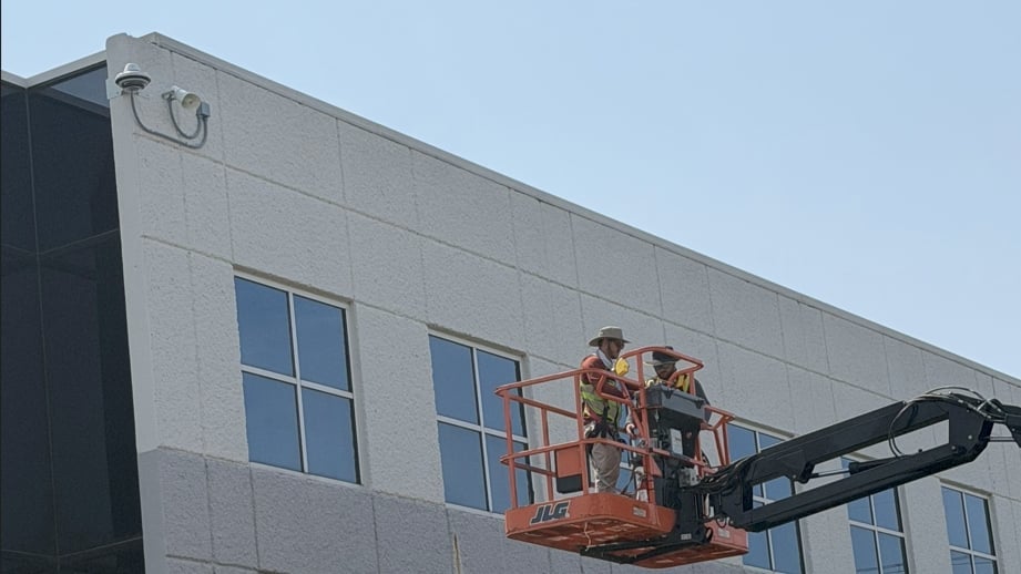 Two workers on a red hydraulic lift platform performing maintenance on the exterior of a modern gray building with large windows.