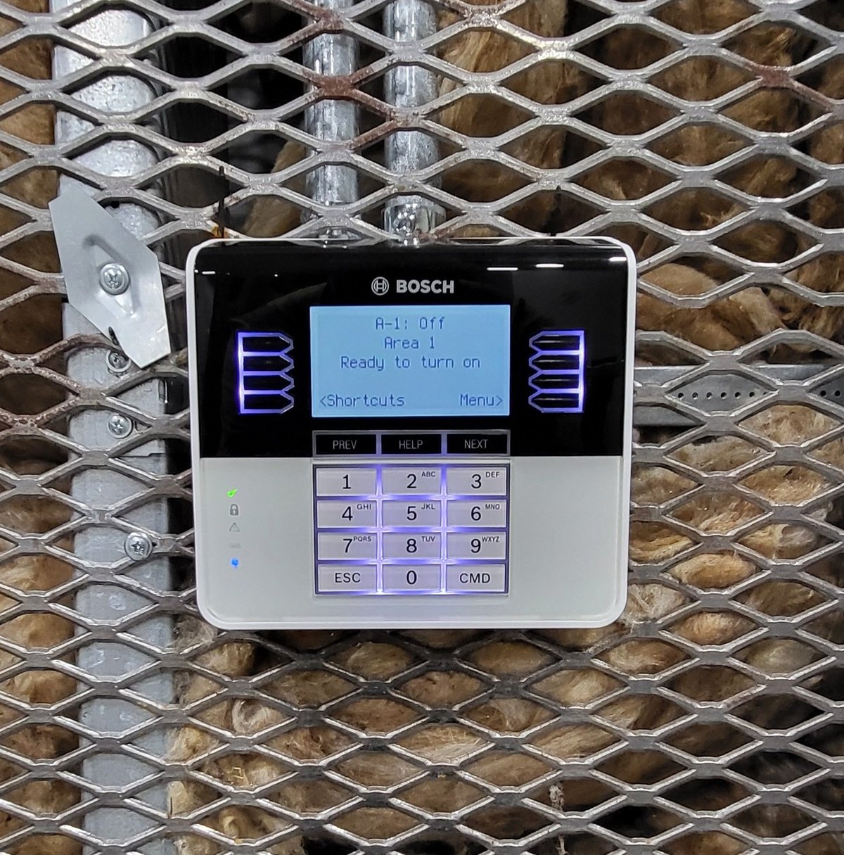 Bosch security alarm keypad mounted on a metal mesh wall, displaying blue LCD screen with Ready to Arm on message