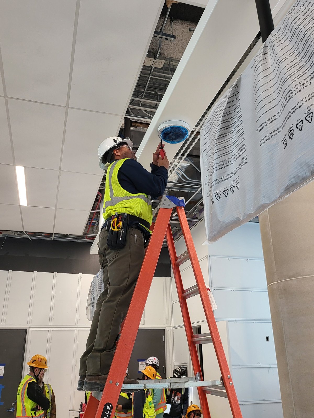 Technician in a yellow safety vest on a red A-frame ladder installing a commercial dome security camera to a drop ceiling in a commercial building, with other workers in hard hats visible below
